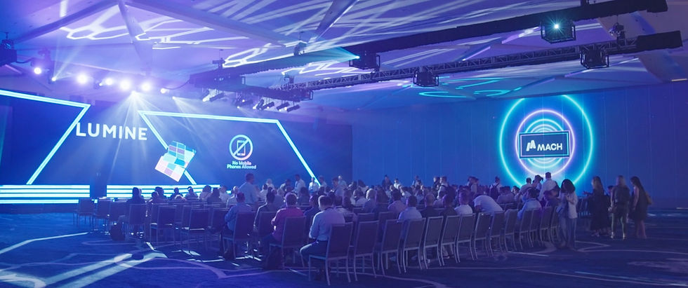 Audience seated in a large, blue-lit conference hall with a stage. Signs read "LUMINE" and "No Mobile Phones Allowed."