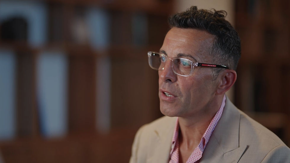 Hombre con gafas claras habla, lleva saco beige y camisa rosa a cuadros. Fondo difuminado de estanterías. Ambiente serio.