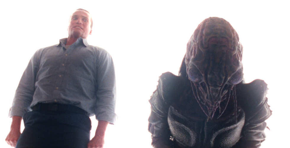 An alien prisoner in human form is bathed in white light with an insect like alien.