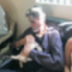 A man with a gray beard and wearing a plaid shirt sits on a couch, gently holding and petting a small, fluffy puppy.
