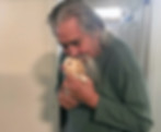 An elderly man with long gray hair and a beard is holding and kissing a small, fluffy puppy.
