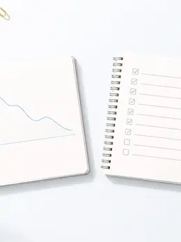 Vue de dessus de deux carnets ouverts posés sur un fond bleu très clair. Le premier montre un graphique en ligne descendant, le second une liste de tâches en deux colonnes avec des cases à cocher, dans une esthétique minimaliste et calme.