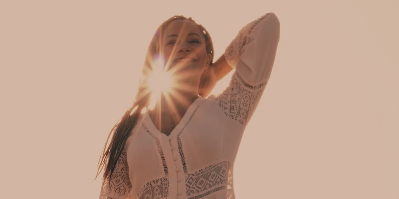 Frau in weißem Spitzenoberteil steht mit geschlossenen Augen im Sonnenlicht, eine Hand im Nacken