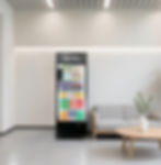 Black 24H Smart Vending machine in a bright modern waiting lounge with gray sofa, round coffee table, and potted plant against a white wall.