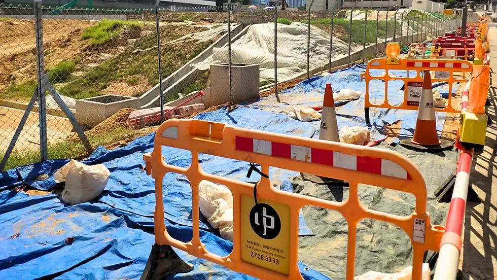Construction site with orange barriers and cones on blue tarp. Fenced-off area, hills in background, caution signs visible.