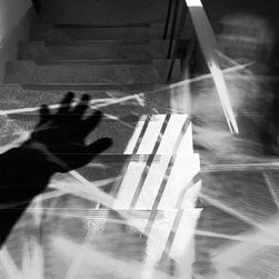 A black-and-white, abstract photo of a staircase with blurred light streaks and a dark hand shadow reaching across the steps. Motion blur suggests a figure moving along the railing, creating a sense of disorientation and movement.