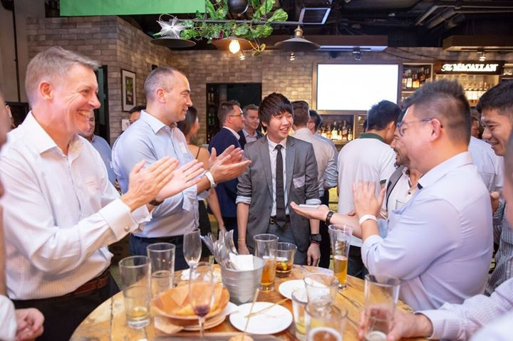 People in business attire are clapping and smiling at a social gathering in a bar with drinks on a wooden table and a lively atmosphere.