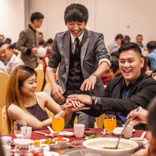 People smiling and laughing at a banquet table with drinks and food. A man in a suit places hands on a woman's hands, creating a joyful mood.