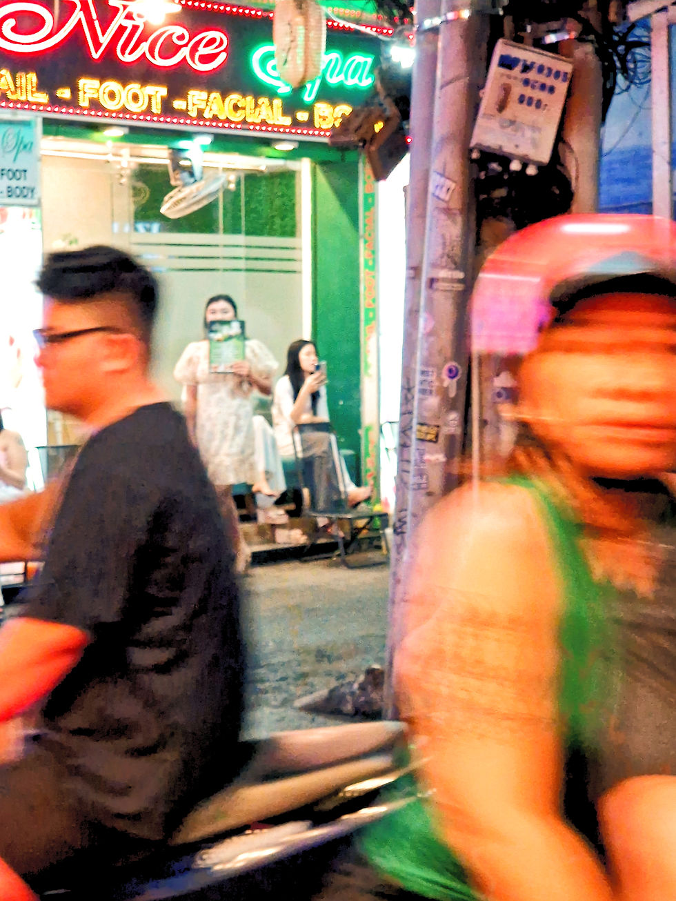 Two moped riders in the streets of Ho Chi Minh city with a beauty parlour in the background and neon lights