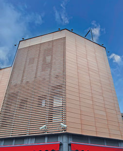 Vista dal basso di una porzione di facciata ventilata in cotto imprunetino con sistema brise-soleil integrato e dettaglio delle tende parasole rosse alla base del centro commerciale.