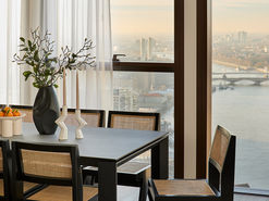 A modern dining area in a Thames City apartment featuring a black rectangular dining table with cane-backed chairs, styled with sculptural white candlesticks and a matte black vase with greenery. Floor-to-ceiling windows reveal panoramic views over the River Thames and London skyline, softly lit by morning light.