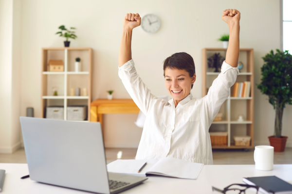 Woman in a white shirt celebrates with raised arms at a desk with a laptop and notebook, in a bright room with shelves and plants.