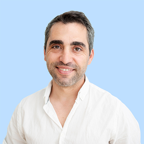 Smiling man in a white shirt with short dark hair and light stubble, posing against a light blue background.