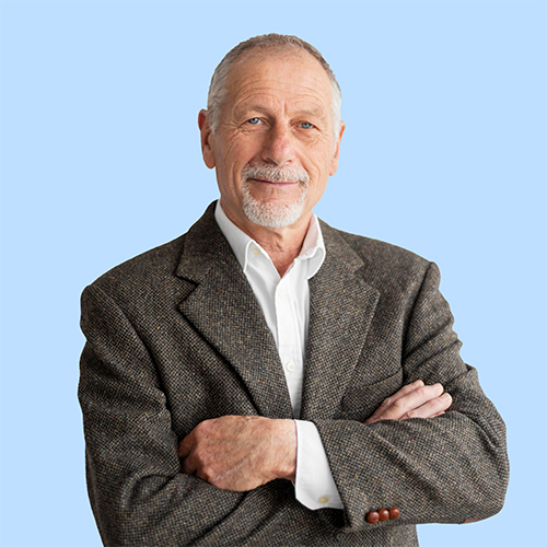 Mature man with a gray goatee and short hair, dressed in a brown blazer and white shirt, standing with arms crossed against a