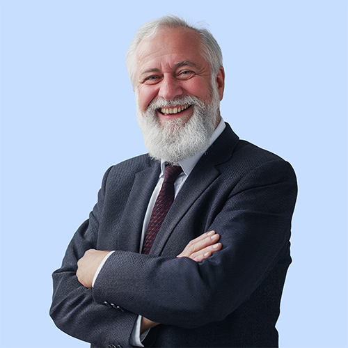 Senior man with a white beard wearing a dark suit and tie, smiling confidently with arms crossed in front of a light blue background.