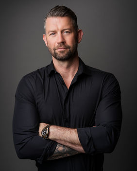 Man in a black button-up shirt, arms crossed, with a confident expression.