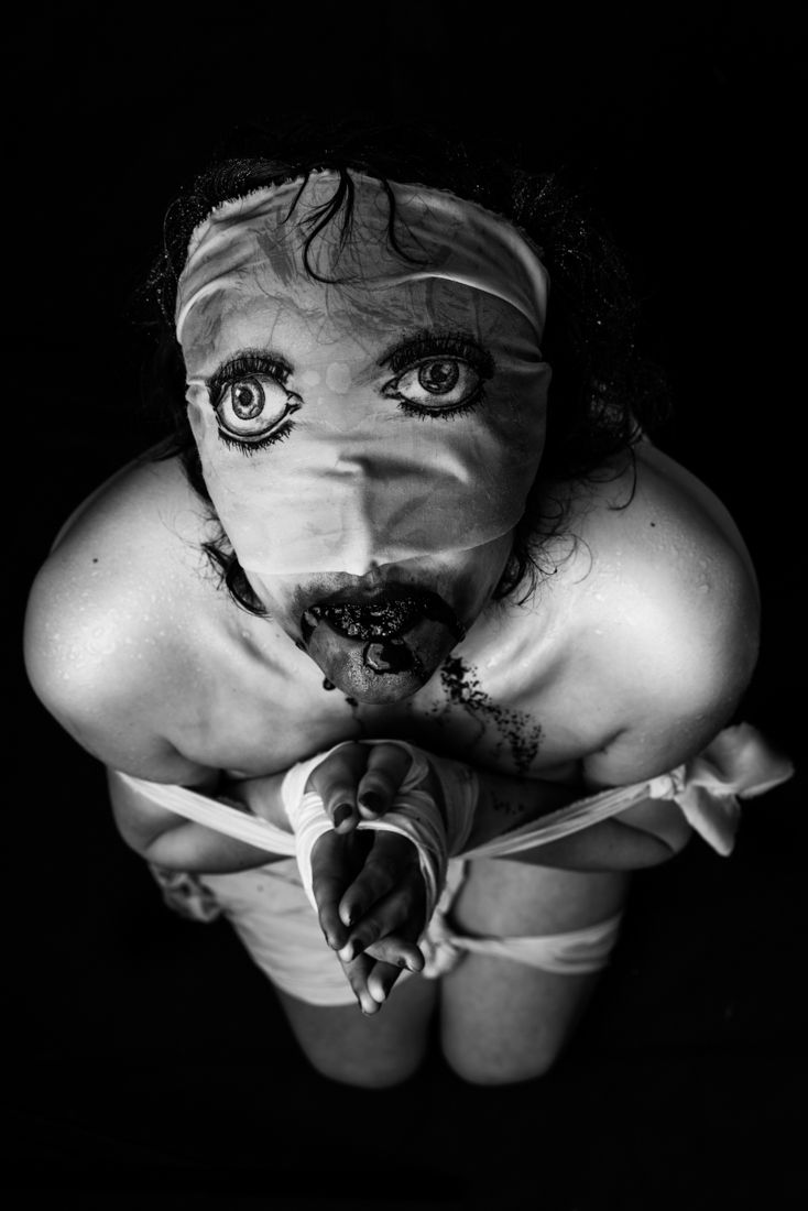 Abstract black and white photo of a woman with drawn-on eyes over a blindfold and dark substance in her mouth, portraying nightmare imagery