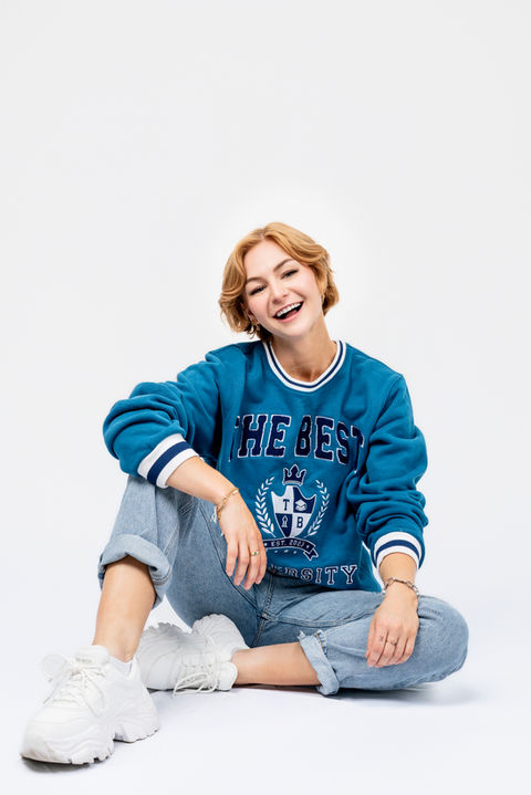 kallmekris sitting cross-legged in Otto blue sweatshirt and jeans, smiling