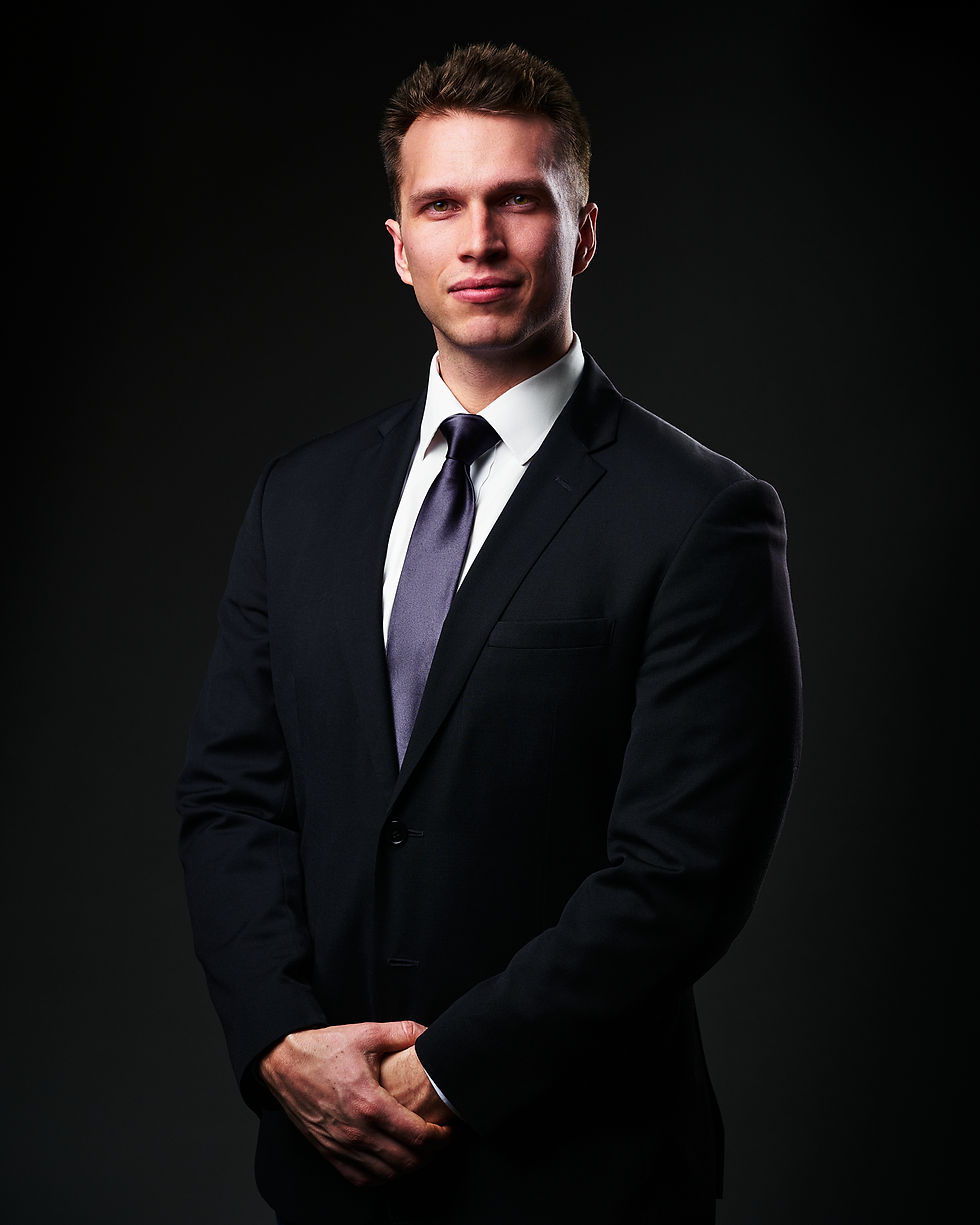 Professional headshot of a man in a dark suit and tie, standing confidently against a dark background with studio lighting.