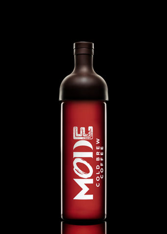 A red bottle labeled 'MODE Cold Brew Coffee' with a dark brown cap, set against a black background. The bottle design is sleek, with bold white text contrasting against the deep red color.