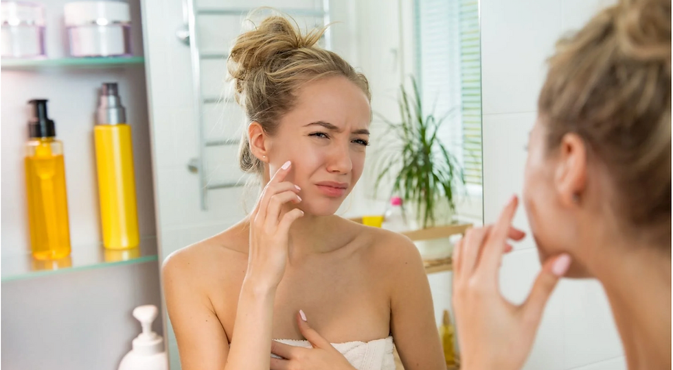 A woman with a towel examines her face in the bathroom mirror, looking concerned. Yellow bottles and a plant are visible in the background.