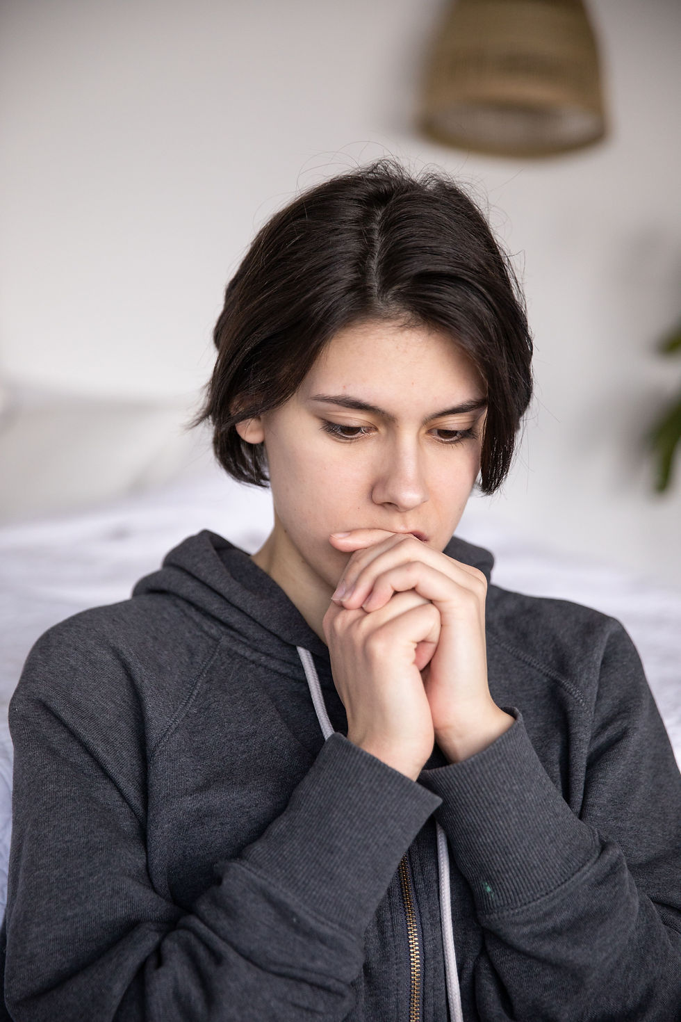 A pensive individual in a hoodie contemplates deeply, resting their chin on clasped hands, while seated in a serene, softly lit room.