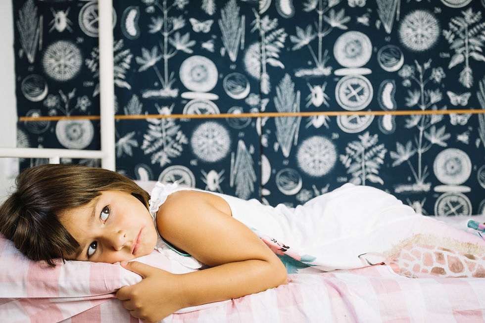 Child in white outfit lying on a pink checkered bed, looking thoughtful. Dark blue wall with botanical patterns in background.