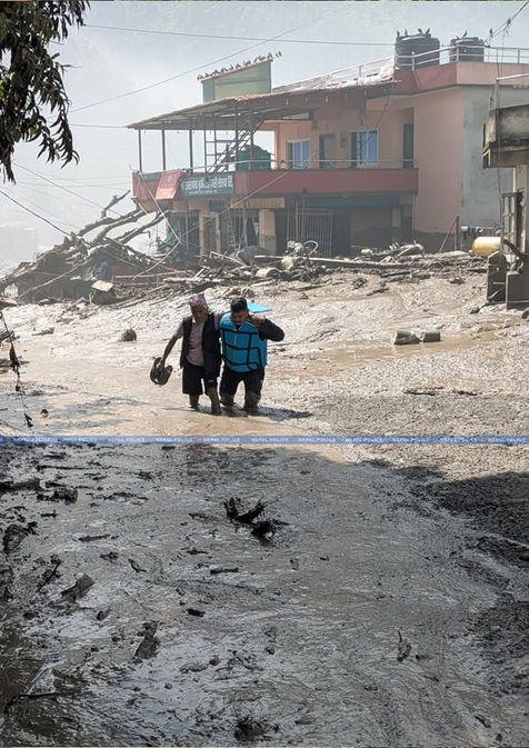 epal Police personnel assisting a person through a flood-damaged area surrounded by mud, debris and damaged buildings in Nepal.