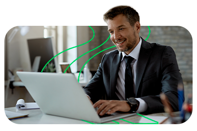 Homem de negócios sorridente, usando terno e blazer escuro, trabalha em um laptop em um escritório moderno, simbolizando sucesso e inovação empresarial.