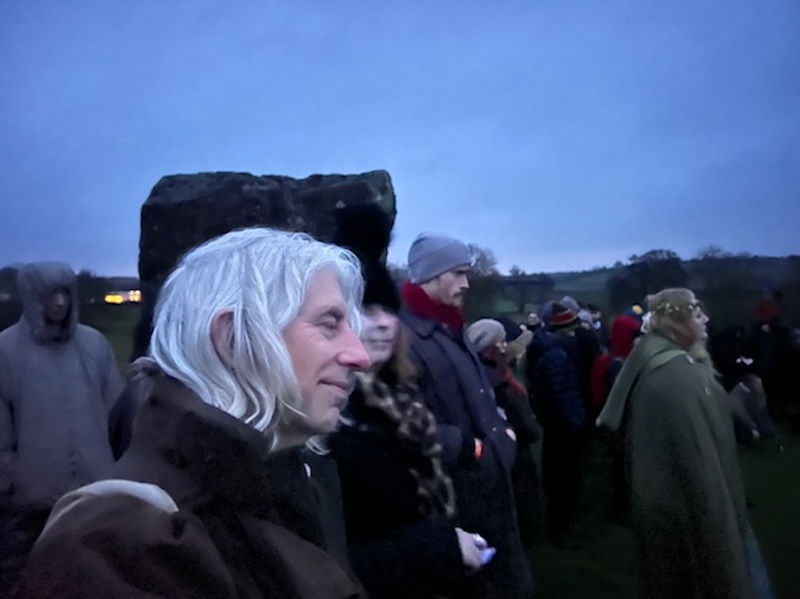 People in winter clothing stand outdoors at dawn, a large stone behind. The scene has a calm, contemplative mood, with a dark sky background.