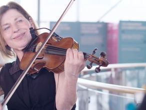 Gina playing violin with a smile in a bright indoor setting. Blurred background with informational panels. Warm, joyful atmosphere.