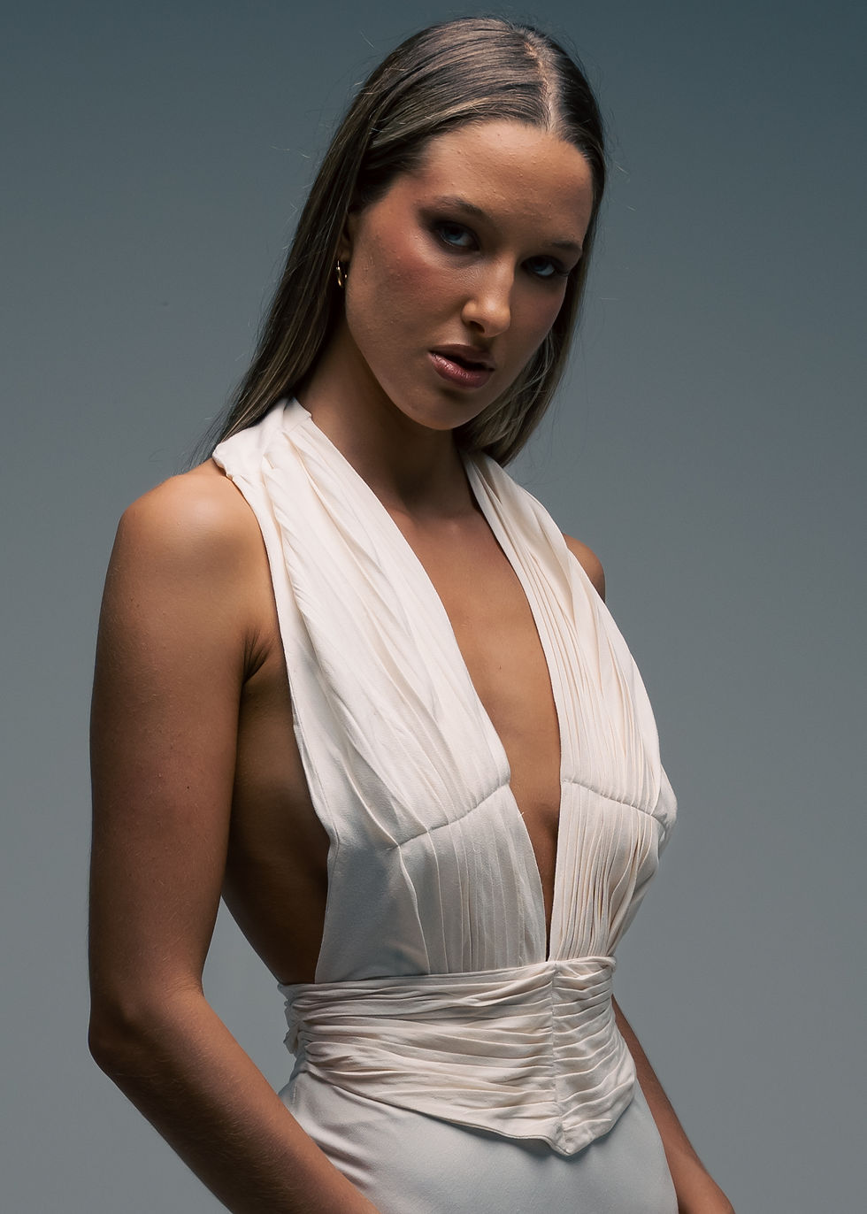 Thumbnail: Close-up of model wearing a cream crepe back satin gown with a deep V neckline, softly pleated bodice and sculpted waistband, captured in soft directional lighting, designed by FLUID the Label Australia