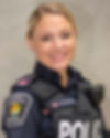 A woman stands before a pale background. She is wearing her police uniform and smiling brightly.