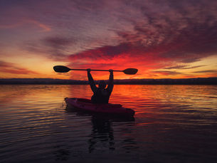 Kayaker raises paddle triumphantly against a vibrant red and purple sunset over calm water, creating a serene and triumphant mood