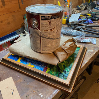 Framed collage lying on workbench, weighted down by gallon can of paint (cushioned with packing paper)