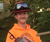 Drew Anderson is a white man with a patchy red beard and mustache and stretched earlobes. He is wearing a neon orange MU Fire and Rescue Training Institute hoodie and a fireperson's hat and goggles.