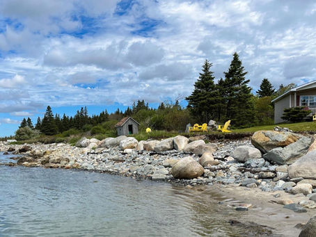Rocky shoreline with yellow chairs near a cottage and small shed, under a partly cloudy sky. Evergreen trees in the background. Peaceful setting.