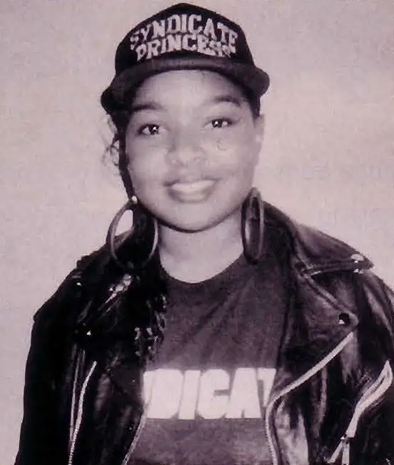 Smiling woman in a Syndicate Princess cap, leather jacket and graphic T-shirt, standing against a plain wall.