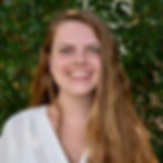 Young woman with long brown hair smiling against green foliage background.