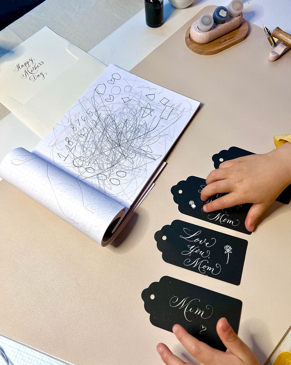 A child’s hands are touching black calligraphy tags with handwritten messages like “Love you, Mom.” Beside them is an open sketchbook filled with childlike scribbles and shapes, next to calligraphy supplies on a soft beige surface.