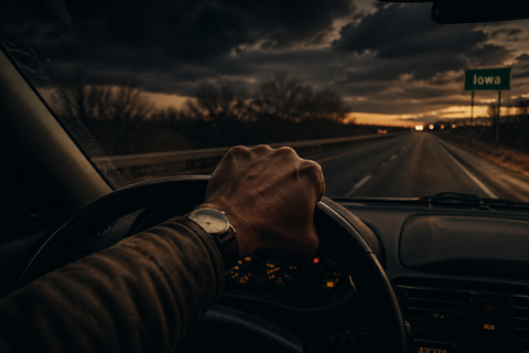 Hand on steering wheel driving toward Iowa at sunset with dark clouds and fading highway lights