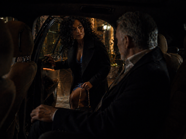Woman pausing at a car door while a man watches from inside, set in a softly lit nighttime scene with quiet tension