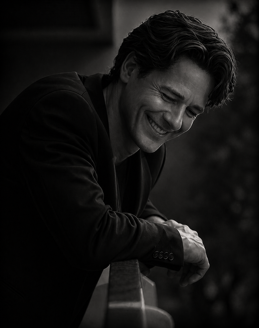 Black and white portrait of Jameson leaning over a railing, head tilted downward with a relaxed smile in a candid, reflective moment.