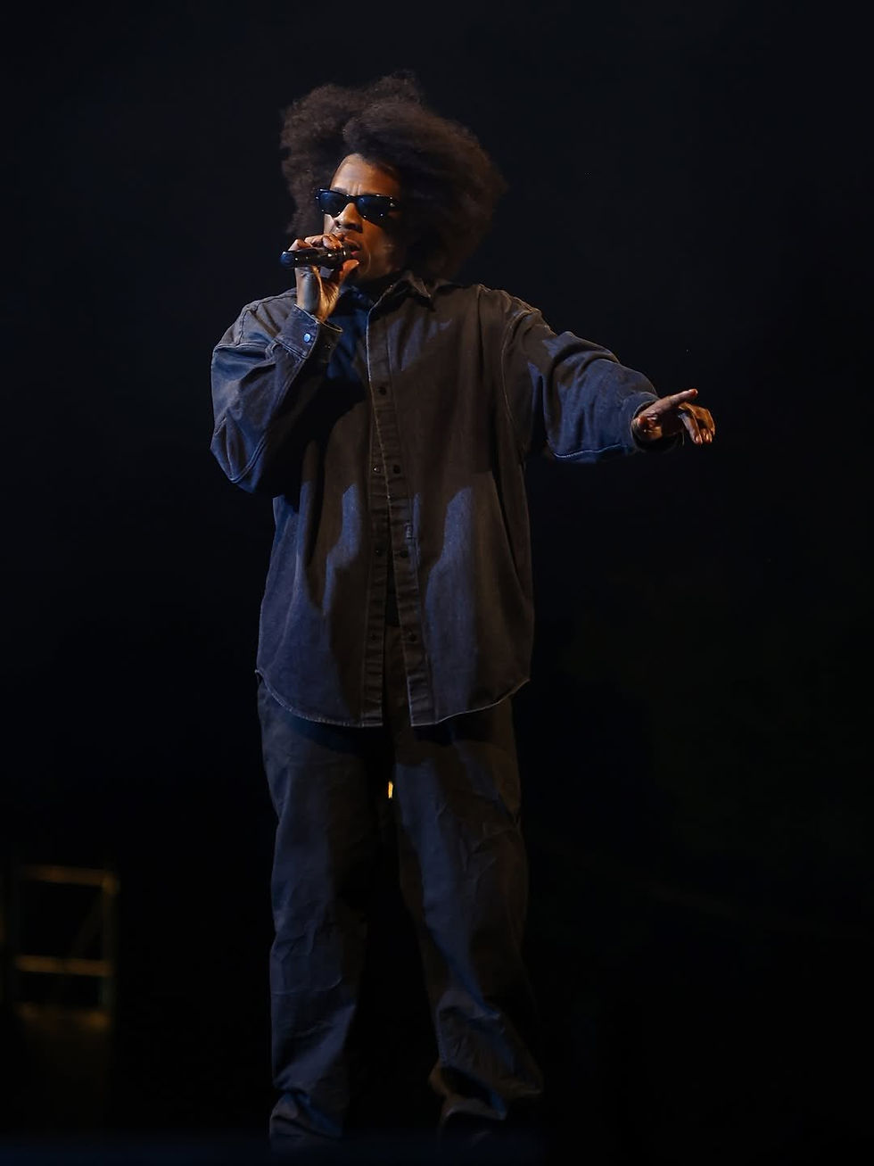 Jay-Z with afro and sunglasses performs on a dark stage, singing into a mic and pointing outward in a cool, focused pose