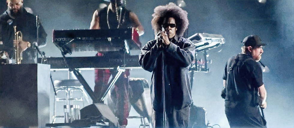 Jay-Z with afro and sunglasses sings into a mic on a smoky stage, backed by band members and keyboards under blue lights.