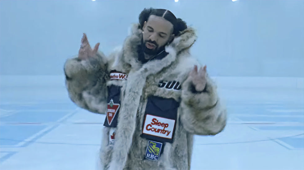 A man, Drake, in a large fur-trimmed coat performs in a cold blue-white room, gesturing with both hands while surrounded by icy lighting and a minimal set.