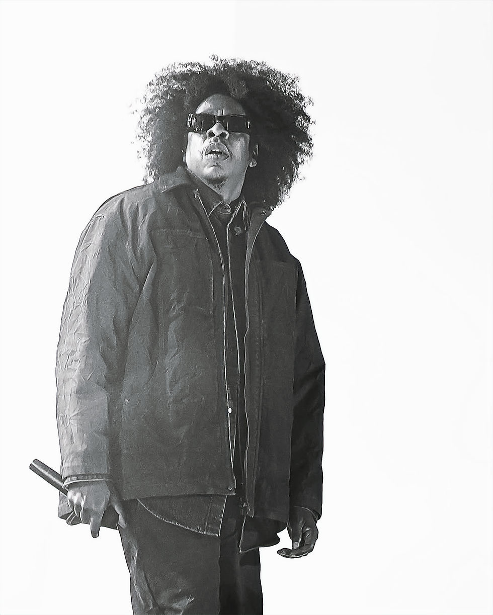 Black-and-white portrait of a Jay-Z with an afro and sunglasses, in a dark jacket, holding a microphone against a plain white background