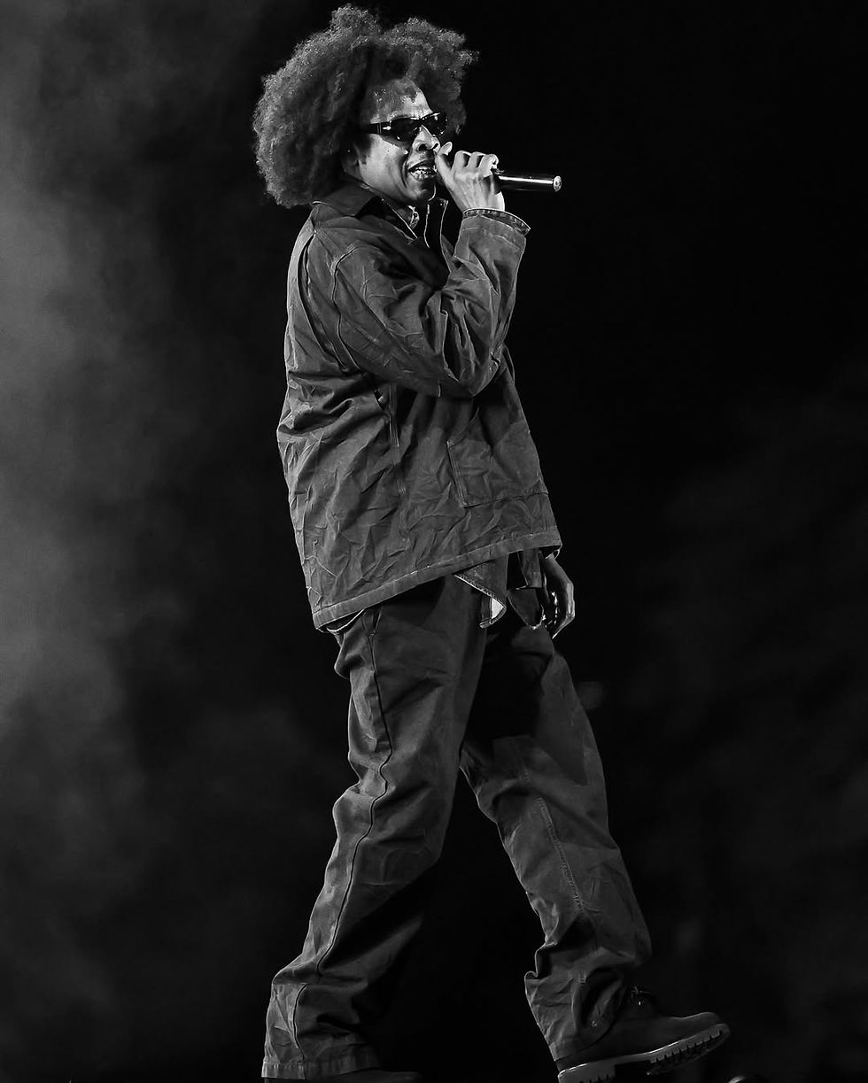 Black-and-white photo of a Jay-Z an afro and sunglasses performing into a microphone on a dark stage.
