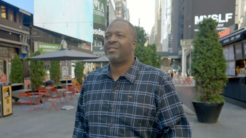 Black man wearing a dark blue plaid shirt stands in Times Square during an outdoor television interview.