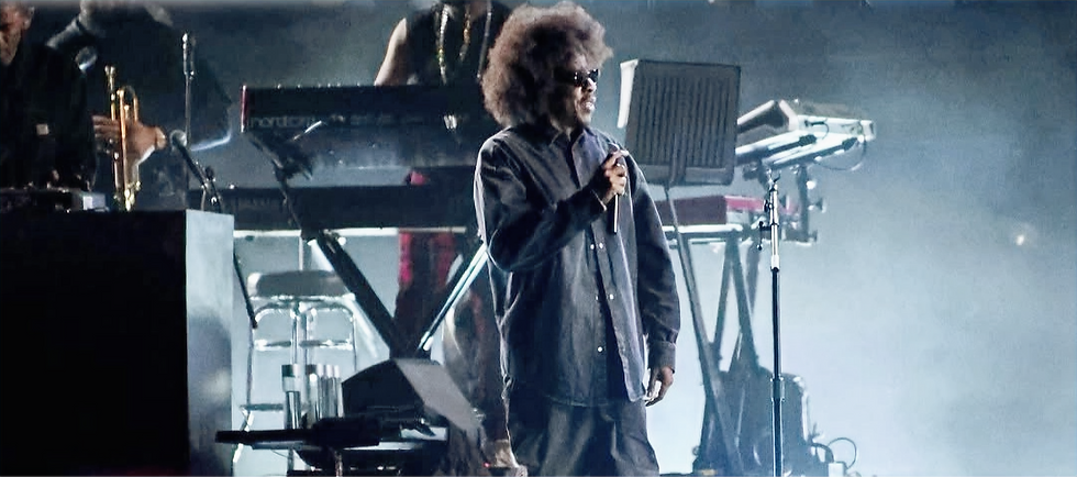 Jay-Z with afro and sunglasses holds a microphone on a smoky stage, with keyboards and band gear behind him.